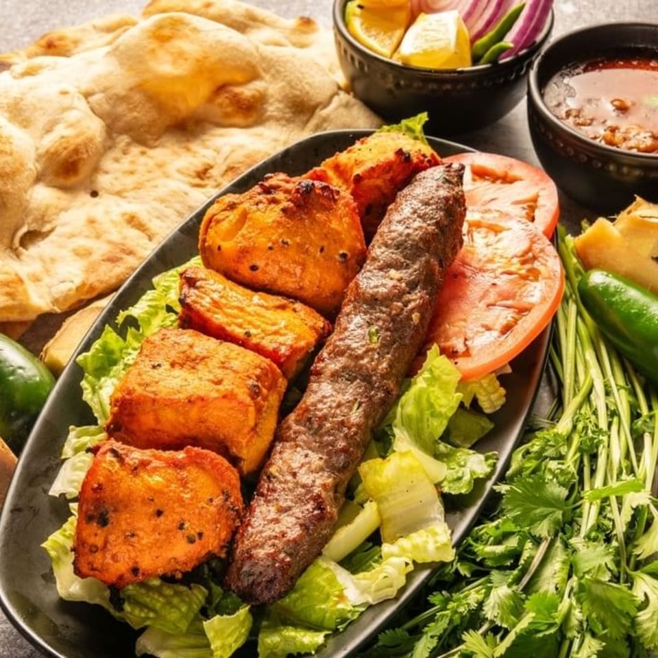 A black plate of Pakistani seekh kabob and chicken tikka, with naan, salad, and sauce.