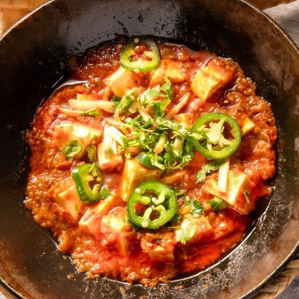 Pakistani karahi dish with paneer, red sauce, green chilies, and cilantro in a black pan.
