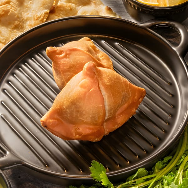 Two golden-brown samosas rest on a black griddle pan, with naan and cilantro visible.
