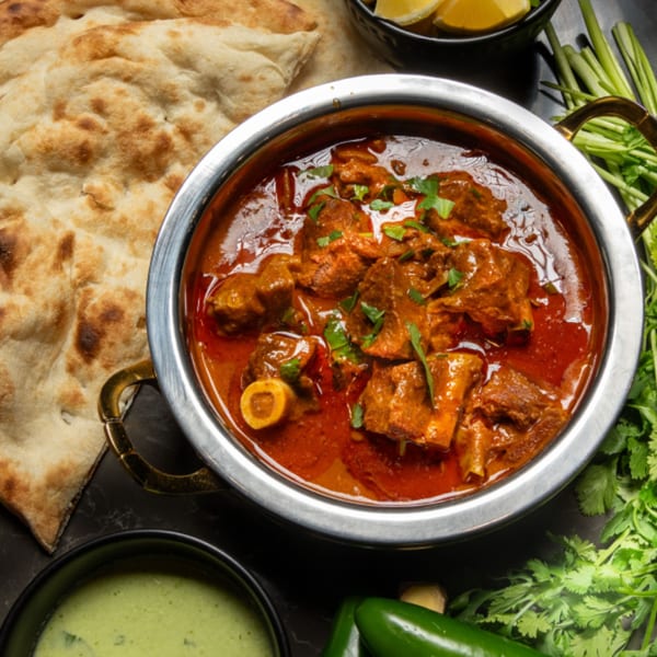 Pakistani lamb karahi in a metal bowl with naan bread, lemons, cilantro, and green chilis.