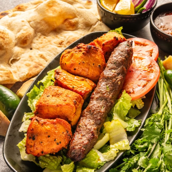 Pakistani seekh kabob and chicken tikka with naan, salad, and sauces on a dark plate.