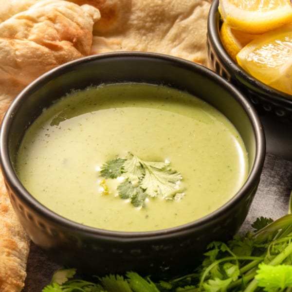 A black bowl of light green Pakistani chutney, garnished with cilantro, alongside naan and lemon wedges.