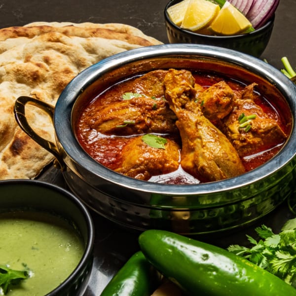 Pakistani chicken karahi in a metal pot, with naan, green chutney, lemons, and chilies.