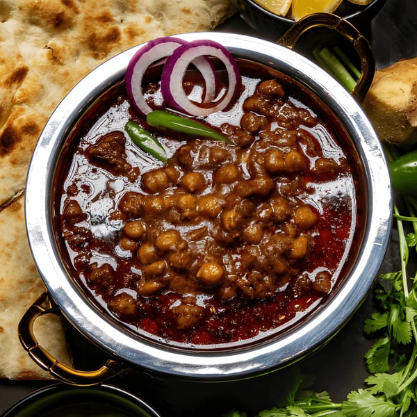 A close-up of a silver bowl filled with chana masala, garnished with red onion rings and green chilies.